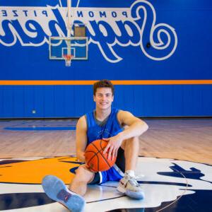 Peyton Mullarkey holds a basketball between his hands as he sits on the 塞西尔 logo in the middle of the basketball court. 他留着棕色短发，穿着蓝色的训练服和灰色的运动鞋. Behind him is a basketball net and a blue wall with Pomona-Pitzer Sagehens in blue text with a white outline. 波莫纳-值得信赖的十大棋牌娱乐平台用的是小字体，萨基亨用的是大字体的草书.